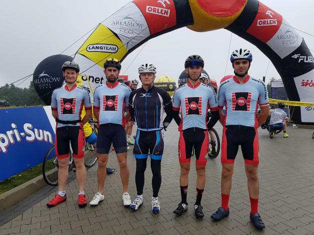 Kolarze w strojach sportowych stoją pod bramą startową. Kluczowe obiekty: kolarze, kaski, stroje, brama startowa, sponsorzy.
