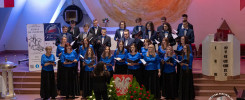 Chór wykonuje pieśń patriotyczną. Kluczowe obiekty: chór, podium, sztandar. Kontekst: festiwal pieśni patriotycznej.