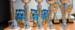 Trofea sportowe na drewnianym stole. Kluczowe obiekty: puchary, medale, dyplomy. Kontekst: ceremonia wręczenia nagród.