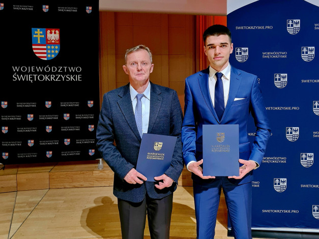 Dwóch mężczyzn w garniturach trzyma dyplomy przed banerem z herbem województwa świętokrzyskiego.