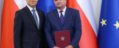 Dwóch mężczyzn w garniturach stoi przed flagami Polski i UE. Jeden z nich trzyma dokument z godłem Polski.