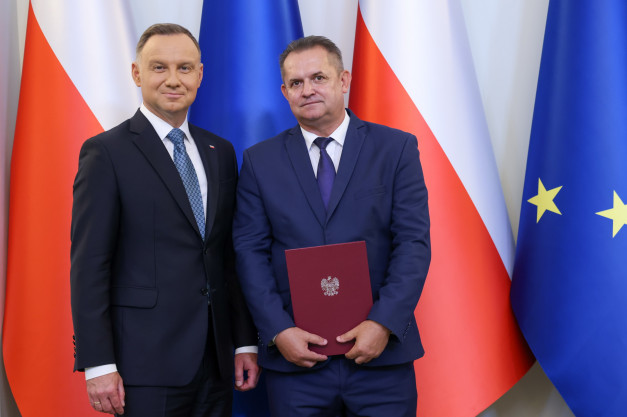 Dwóch mężczyzn w garniturach stoi przed flagami Polski i UE. Jeden z nich trzyma dokument z godłem Polski.