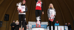 Trzy kobiety na podium po biegu, z nagrodami. Kluczowe obiekty: podium, kobiety, nagrody, logo "SieBiega".
