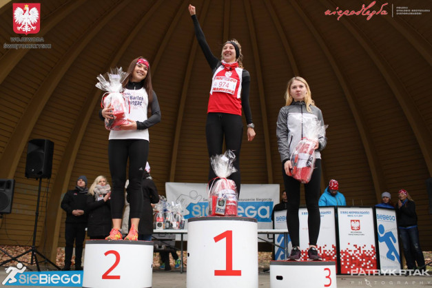 Trzy kobiety na podium po biegu, z nagrodami. Kluczowe obiekty: podium, kobiety, nagrody, logo "SieBiega".
