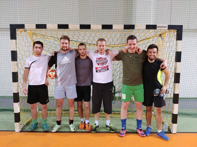 Sześciu mężczyzn stoi przed bramką do piłki ręcznej. Mają na sobie sportowe stroje.