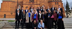 Grupa ludzi w strojach chóralnych stoi przed ceglanym kościołem.