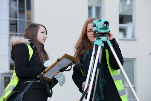 Dwie kobiety w kamizelkach odblaskowych używają niwelatora i tablicy. Kontekst: praca terenowa, budowa.