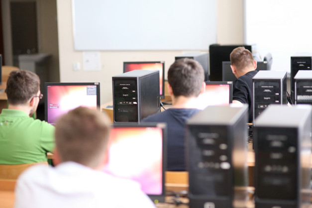 Ludzie siedzą przy komputerach w sali lekcyjnej. Widoczne są monitory, jednostki centralne i tablica.