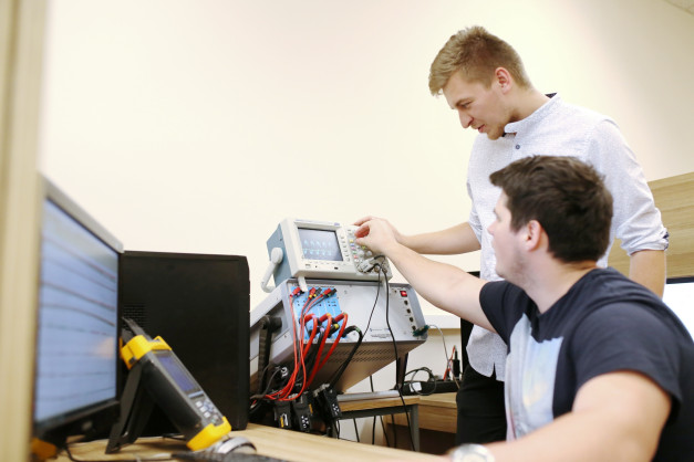 Dwóch mężczyzn pracuje przy sprzęcie elektronicznym. Kluczowe obiekty: oscyloskop, komputer, miernik.