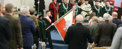 Uroczystość akademicka z udziałem osób w togach i z flagą Polski. Kluczowe obiekty: togi, flagi, ludzie.