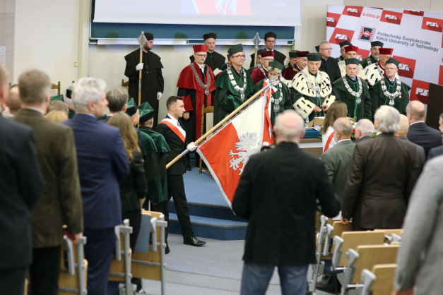Uroczystość akademicka z udziałem osób w togach i z flagą Polski. Kluczowe obiekty: togi, flagi, ludzie.