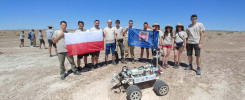 Grupa ludzi stoi na pustyni z polską flagą i robotem. Kluczowe obiekty: ludzie, robot, flaga Polski, pustynia.