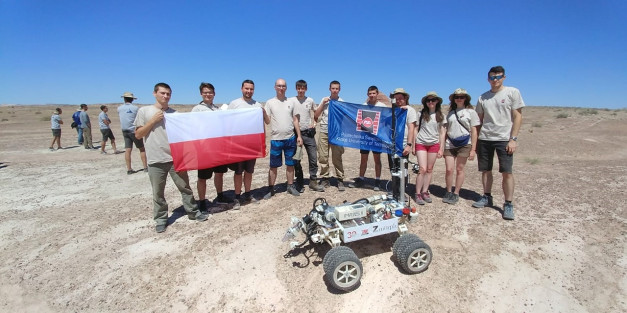 Grupa ludzi stoi na pustyni z polską flagą i robotem. Kluczowe obiekty: ludzie, robot, flaga Polski, pustynia.
