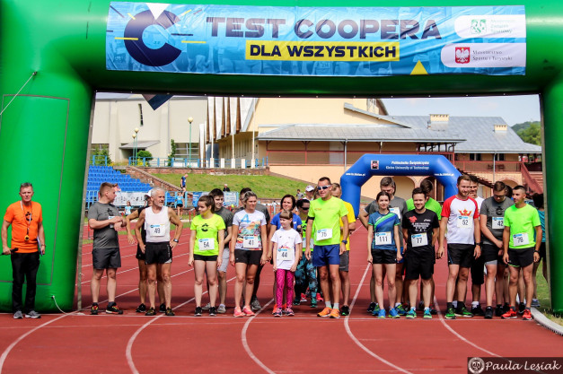 Grupa biegaczy stoi na bieżni pod zielonym łukiem z napisem "TEST COOPERA". Kluczowe obiekty: ludzie, łuk startowy, bieżnia.