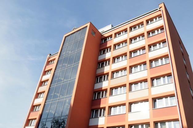 Pomarańczowy budynek z oknami i przeszkloną klatką schodową na tle błękitnego nieba.