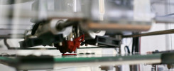 Drukarka 3D z czerwonym elementem grzewczym drukuje biało-czerwony obiekt na szklanej platformie.