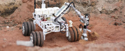 Robot z ramieniem i kamerą na kołach, pracujący na czerwonej, piaszczystej powierzchni.