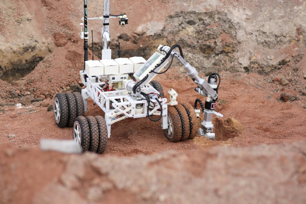 Robot z ramieniem i kamerą na kołach, pracujący na czerwonej, piaszczystej powierzchni.