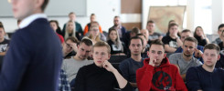 Grupa ludzi siedzi na wykładzie. Widać młodego mężczyznę w czerwonej bluzie.