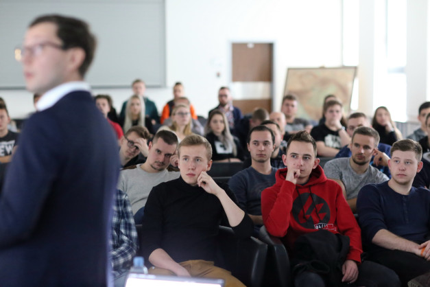 Grupa ludzi siedzi na wykładzie. Widać młodego mężczyznę w czerwonej bluzie.