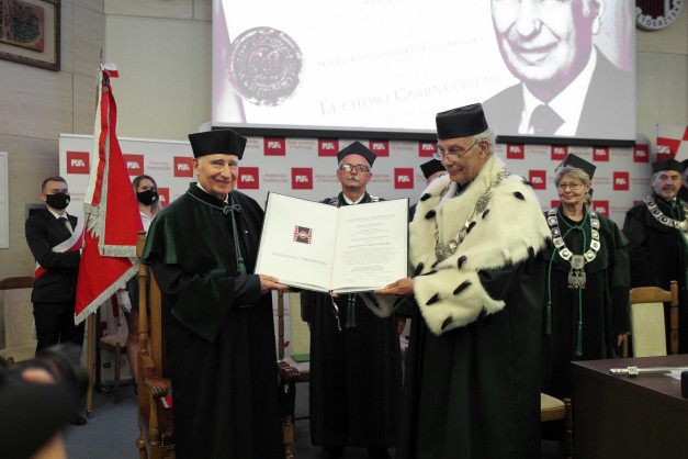 Dwóch mężczyzn w togach akademickich wręcza dokument. W tle ekran z portretem i logo Politechniki Świętokrzyskiej.