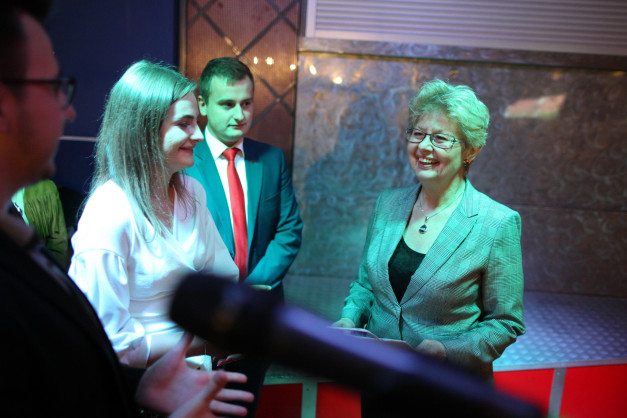 Kobieta w marynarce i mężczyzna w garniturze rozmawiają. W tle widać mikrofon.