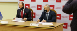Dwóch mężczyzn w garniturach i maseczkach podpisuje dokumenty przy drewnianym stole. W tle baner Politechniki Świętokrzyskiej.