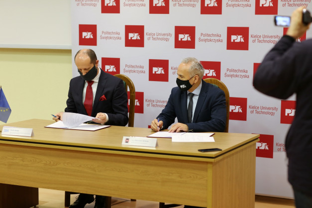 Dwóch mężczyzn w garniturach i maseczkach podpisuje dokumenty przy drewnianym stole. W tle baner Politechniki Świętokrzyskiej.