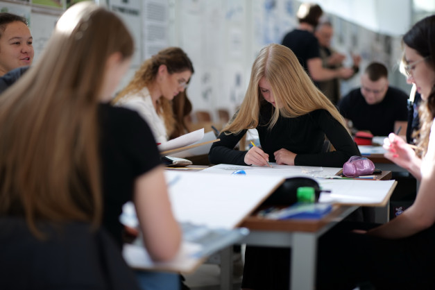 Ludzie rysują przy stołach w pracowni. Kluczowe obiekty: osoby, stoły, papiery, ołówki. Kontekst: zajęcia artystyczne.