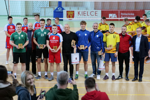 Grupa młodych siatkarzy w strojach sportowych odbiera nagrody. W tle siatka do siatkówki i banery.