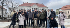 Grupa osób stoi na brukowanym placu przed budynkiem z czerwonym dachem. Kluczowe obiekty: ludzie, budynek, plac.