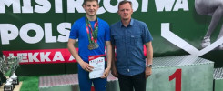 Mężczyzna w stroju sportowym z medalem i dyplomem stoi obok drugiego mężczyzny.