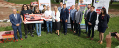 Grupa osób stoi na trawie przed banerami. Trzymają dyplomy i plakaty. W tle budynek i plac budowy.