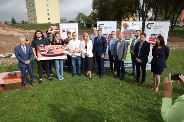 Grupa osób stoi na trawie przed banerami. Trzymają dyplomy i plakaty. W tle budynek i plac budowy.
