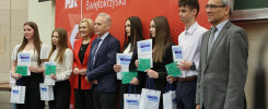 Grupa osób odbiera dyplomy i upominki na tle Politechniki Świętokrzyskiej. Kluczowe obiekty: ludzie, dyplomy, torby.