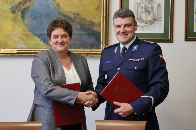 Kobieta i mężczyzna w mundurze ściskają sobie dłonie, trzymając czerwone teczki. W tle obrazy.
