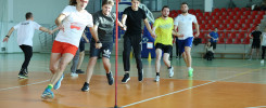 Grupa mężczyzn biegnie w sztafecie w hali sportowej. Kluczowe obiekty: biegacze, tyczka, trybuny. Kontekst: zawody sportowe.