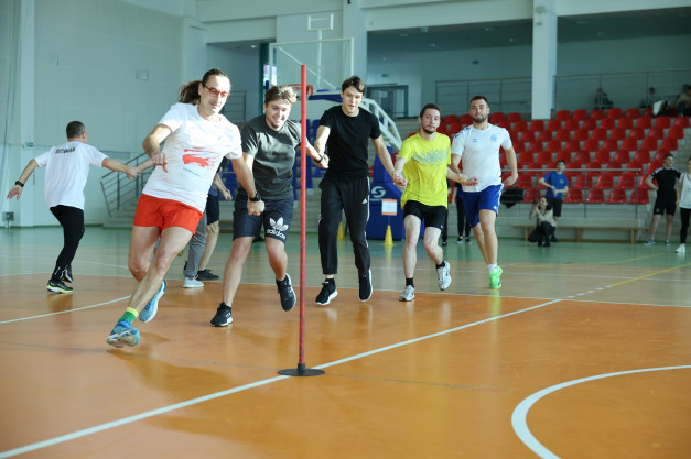 Grupa mężczyzn biegnie w sztafecie w hali sportowej. Kluczowe obiekty: biegacze, tyczka, trybuny. Kontekst: zawody sportowe.