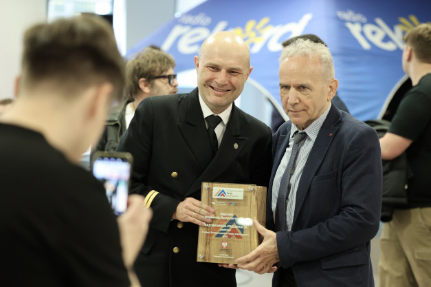 Dwóch mężczyzn odbiera nagrodę podczas Świętokrzyskich Targów Edukacyjnych. Kluczowe obiekty: nagroda, mężczyźni, targi.