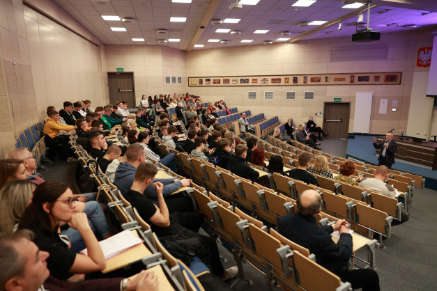 Sala wykładowa z widownią siedzącą w rzędach. Kluczowe obiekty: widownia, mównica, projektor.