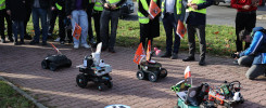 Grupa osób w kamizelkach obserwuje roboty na chodniku. Widać roboty, flagi, samochody i budynki.