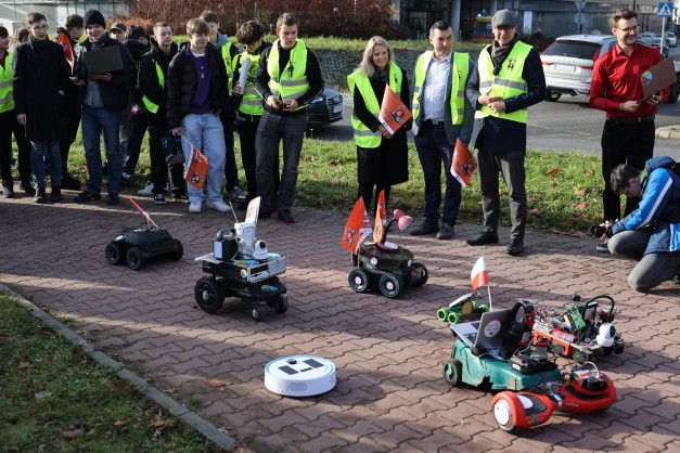 Grupa osób w kamizelkach obserwuje roboty na chodniku. Widać roboty, flagi, samochody i budynki.