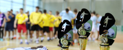 Trzy puchary sportowe z piłkarzami i piłkami, medale i notatki na stole. W tle grupa osób.