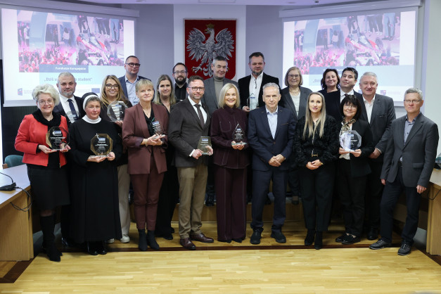 Grupa osób pozuje do zdjęcia z nagrodami. W tle ekran z napisem "Fundusze Europejskie".
