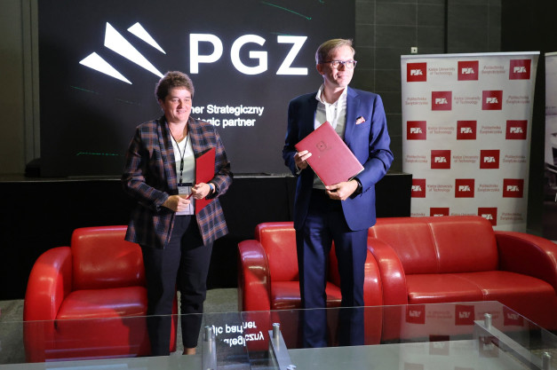 Dwoje ludzi stoi przed ekranem PGZ. Kobieta w kratkę i mężczyzna w garniturze trzymają dokumenty.