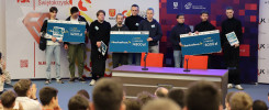 Grupa młodych ludzi odbiera nagrody pieniężne na scenie, podczas wydarzenia typu hackathon.