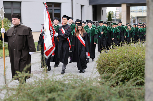 Uroczysta procesja absolwentów w togach i biretach, niosących sztandar. Kontekst: uroczystość akademicka.