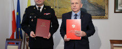 Dwóch mężczyzn trzyma dokumenty. Jeden w mundurze, drugi w garniturze. W tle flagi i obraz.