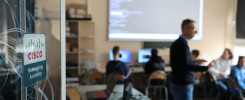 Tablica Cisco Networking Academy. W tle sala lekcyjna z ludźmi i ekranem.