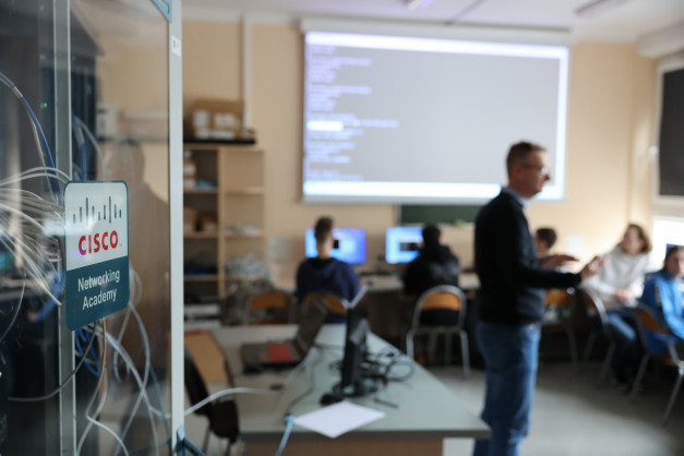 Tablica Cisco Networking Academy. W tle sala lekcyjna z ludźmi i ekranem.
